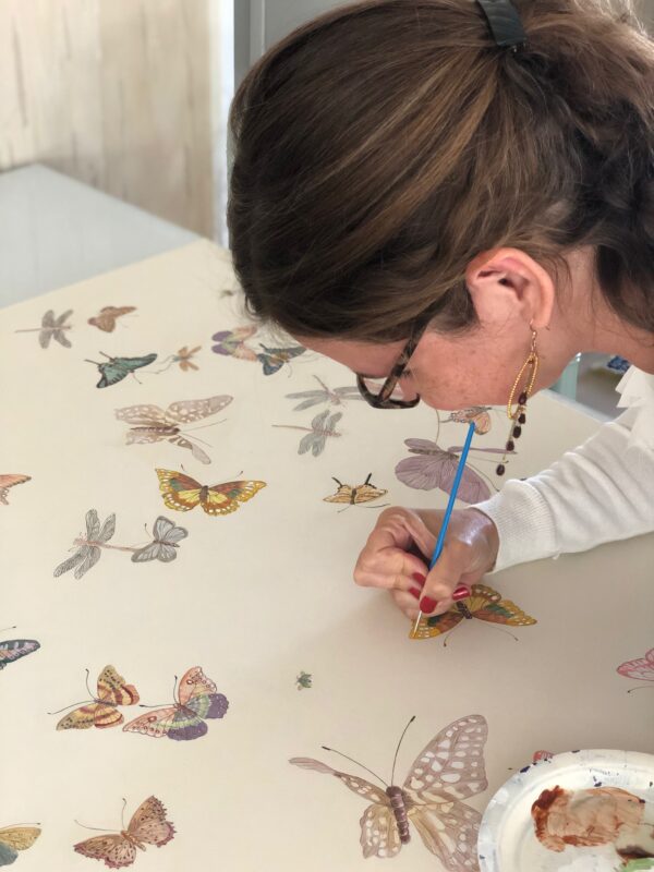 A woman is painting "Daydream" Chinoiserie butterflies on a table.